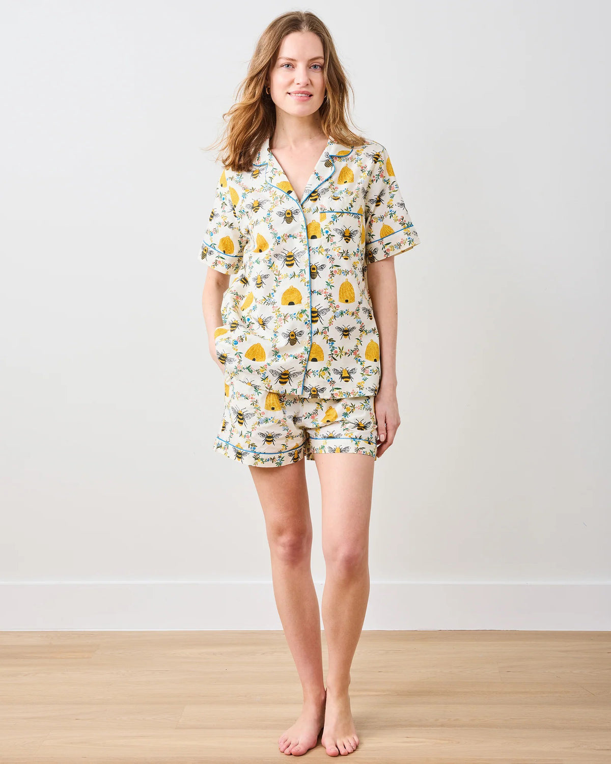 A person with long hair is wearing the PRINTFRESH - SWEET BEE COTTON SHORT SET, featuring a floral pattern in yellow and green. The short-sleeved shirt and shorts exude comfort and style as they stand barefoot on a wooden floor against a plain white wall.