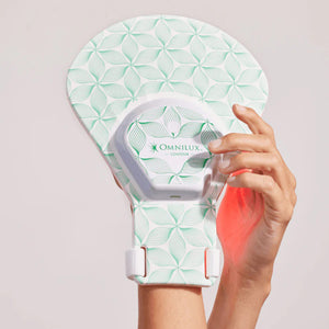 The OMNILUX - COUNTOUR GLOVE, designed by OMNILUX, features a geometric pattern and emits a pink glow on white to highlight its red light therapy benefits for skin rejuvenation.
