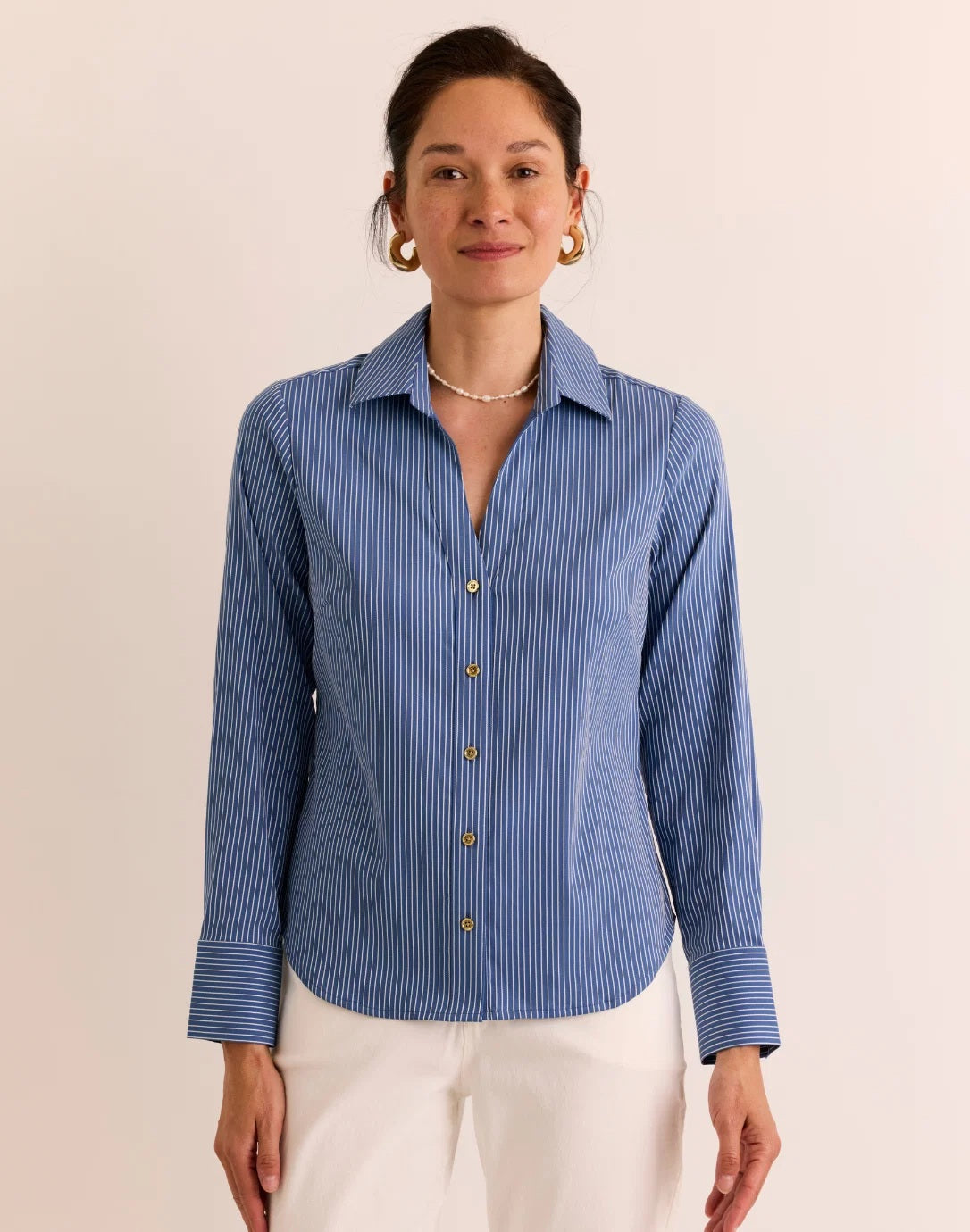 A woman stands against a plain, light background, exuding effortless style and breathable comfort in the HINSON WU Lindsey Long Sleeve Ribbon Stripe Shirt in Blue and White—a lightweight pinstripe button-up featuring gold buttons—paired with white pants.