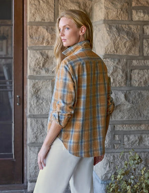 A woman stands indoors against a neutral background with soft light streaming in, smiling gently with one hand in her pocket. She wears the FRANK & EILEEN Eileen Relaxed Button Up Shirt in Italian Flannel Gold Blue Large Plaid—featuring a bust-flattering button placement—paired with cream high-waisted pants and brown ankle boots.