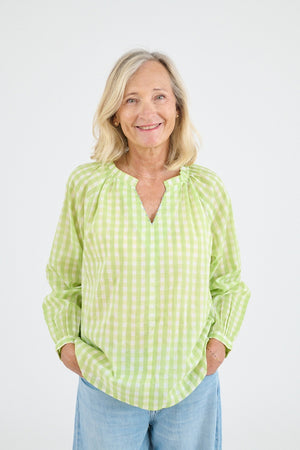 A person with shoulder-length blonde hair, seen from behind, wears the STORIATIPIC IRIS GINGHAM BLOUSE IN GREEN—featuring a light green and white check pattern and embroidered collar—with light blue jeans against a plain white background.