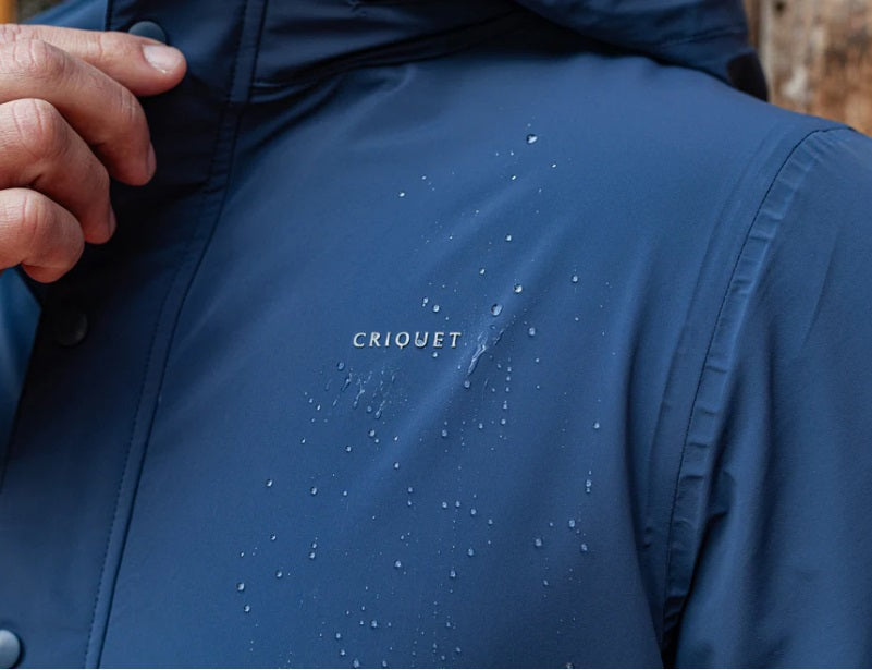 A man wearing the CRIQUET - Watershed Rain Jacket in Navy stands outdoors on a rainy day, with trees and wet pavement in the background.