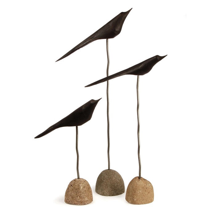 The NAPA HOME AND GARDEN &quot;THE FLOCK&quot; Set of 3-Black minimalist bird sculptures are displayed indoors on slender stands with stone bases, set on a wooden floor against a white brick wall with leafy branches above.