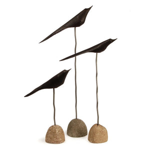 The NAPA HOME AND GARDEN "THE FLOCK" Set of 3-Black minimalist bird sculptures are displayed indoors on slender stands with stone bases, set on a wooden floor against a white brick wall with leafy branches above.