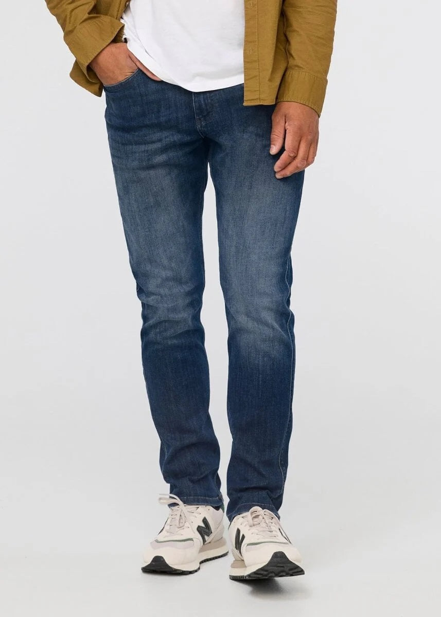 A person wearing a white t-shirt, DUER PERFORMANCE DENIM ATHLETIC TAPER PANTS IN GALATIC, and white sneakers stands facing away from the camera against a plain light background.
