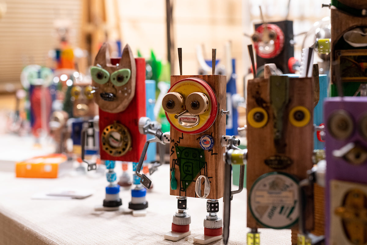 A group of colorful, whimsical robot sculptures made from recycled materials like wood, bottle caps, and metal parts stand on display on a table.