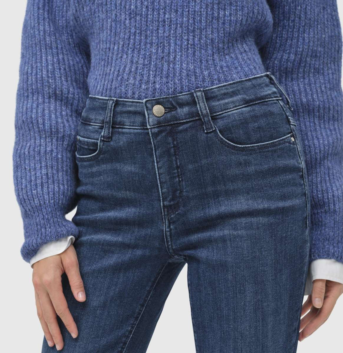 A person stands wearing a blue sweater, white shirt with a bow, and MAC&#39;s DREAM BOOT AUTHENTIC DENIM jeans. They have short light brown hair and pose against a plain light gray background.