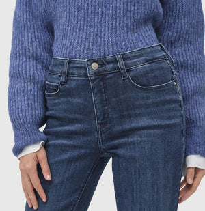 A person stands wearing a blue sweater, white shirt with a bow, and MAC's DREAM BOOT AUTHENTIC DENIM jeans. They have short light brown hair and pose against a plain light gray background.