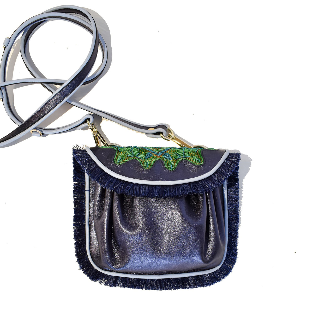 The MEHER KAKALIA Mini Accordion Bag with Fringe Sonic Saphire Tropical is a handcrafted shoulder accessory in shiny dark blue featuring fringe trim and green embroidery on the flap. Its long strap, gold hardware, and elegant design make it a versatile addition to any outfit.