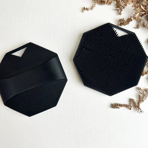 The SILICONE BODY AND EXFOLIATION BRUSH IN ONYX from SMILE BOUTIQUES features a black, octagonal silicone design with a textured, raised dot surface and a small triangular handle cutout at the top—perfect for use as a reusable shower brush or exfoliating body scrubber.