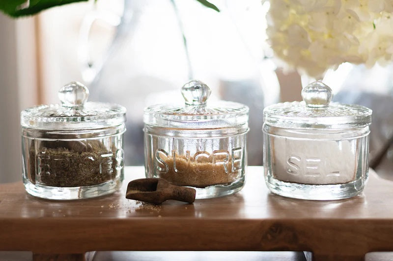 The ETUHOME Bistro Glass Salt Cellar with Scoop sits on a wooden tray atop a marble counter, accompanied by two other glass jars for herbs and sugar. A small wooden scoop from ETUHOME rests in front.