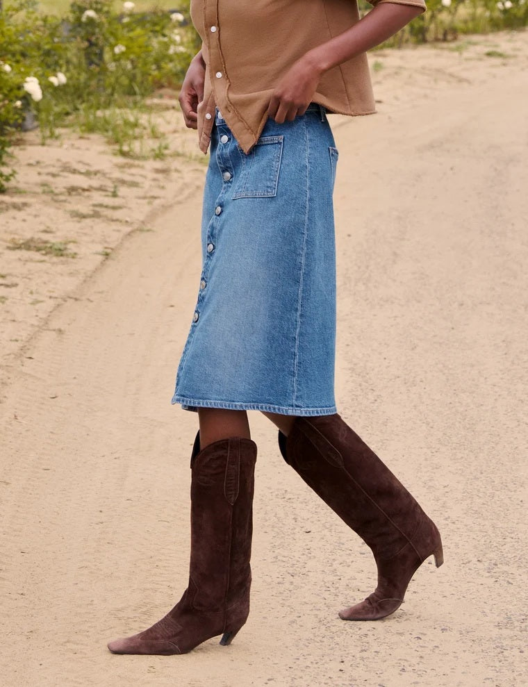 A woman walks on a dirt road near a fence, wearing a tan button-up shirt and the FRANK & EILEEN - SHANNON CLASSIC DENIM SKIRT IN 1978 WASH, paired with brown boots. In the background, trees and horses stand in a grassy field.