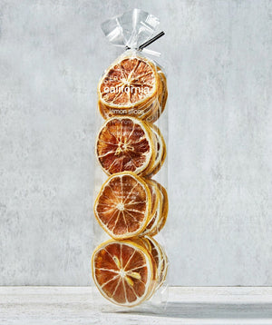 A transparent, tall bag containing neatly stacked dried orange slices. The bag is tied at the top with a clear ribbon. The product label reads "DARDIMANS - CRISPY ORANGE SLICES," hinting at the fresh orange flavor packed with vitamin C. The background is a light textured surface, giving a rustic appearance.