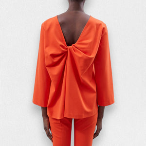A person is wearing the MEIMEIJ - TOP WITH KNOT BACK DETAIL in orange, featuring wide sleeves and a stylish knot back design, paired with matching relaxed-fit orange pants. The outfit appears loose and comfortable, perfect for a casual look. The background is a plain, light-colored texture.