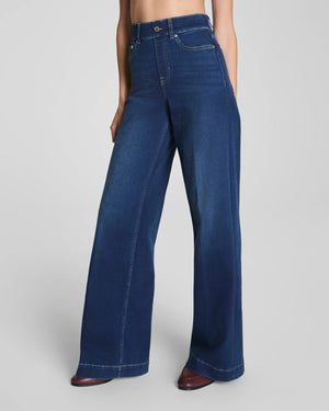 A woman with long brown hair stands against a plain background, smiling slightly. She is wearing a blue and white striped blouse paired with the SPANX Shape EveryWear Pull On Wide Leg Jean in Poseidon by SPANX and brown shoes, accessorized with a watch on her wrist.