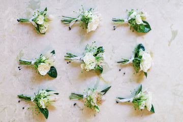 Nine small white floral boutonnieres arranged in a neat three-by-three grid on a light marble surface. Each boutonniere features white flowers, greenery, and is tied with a white ribbon.