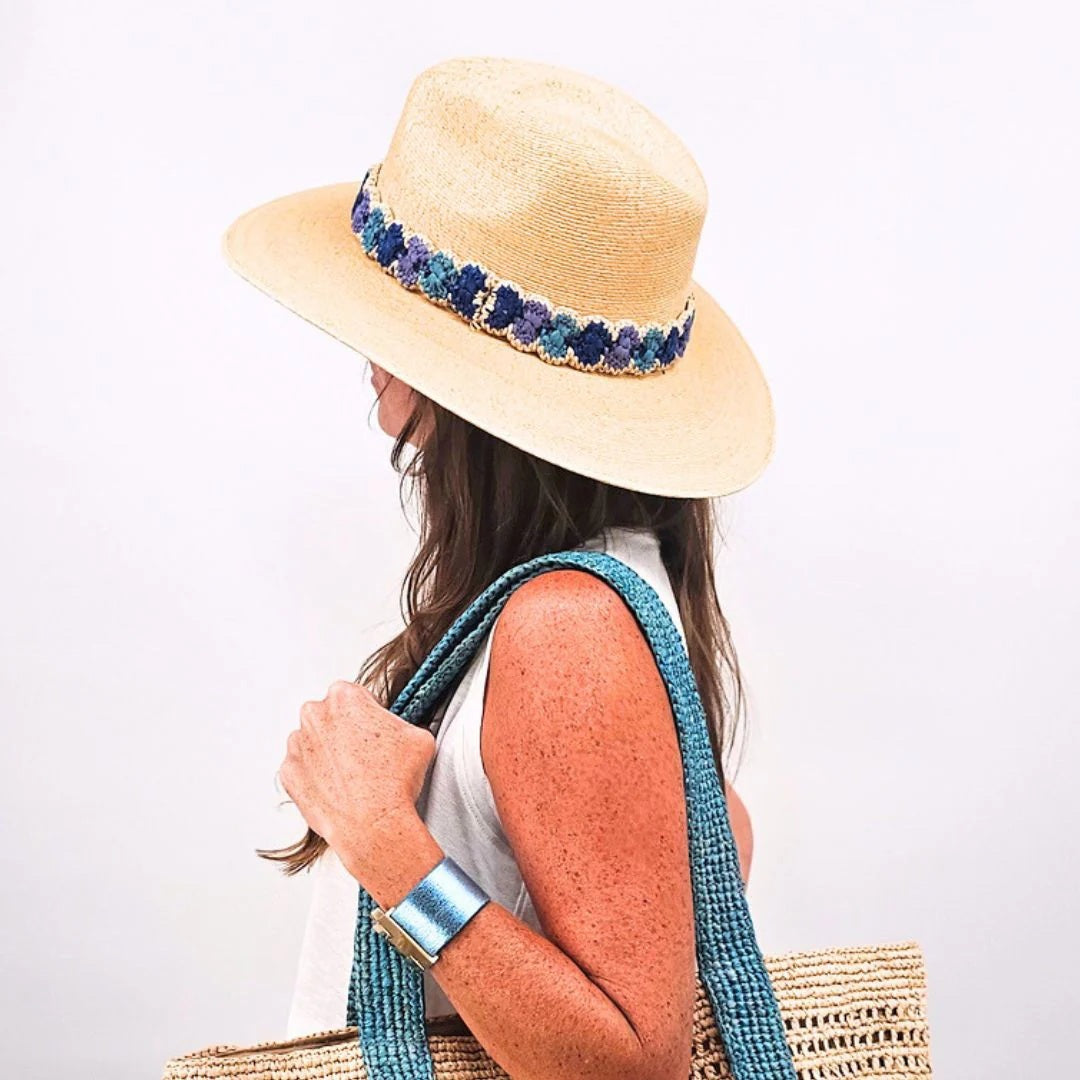 A woman with long brown hair wears the BALDIZ Sand Palm Hat with Blue Ombre Band and holds a woven straw bag with blue handles, plus a metallic blue bracelet. She faces sideways against a white background. One size fits most.