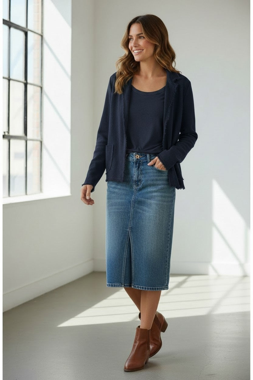A woman smiles as she walks through a bright room with large windows and a gray floor, styled in a navy blazer layered over the BASE MILANO - KNIT COTTON CARDIGAN IN NAVY BLUE, paired with a dark top, blue knee-length denim skirt, and brown ankle boots—a perfect outfit for lightweight layering.