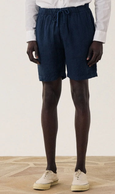 A person wearing a white long-sleeve shirt, HARTFORD - LINEN JOGGY SHORT IN NAVY by HARTFORD, and beige sneakers stands on a wooden floor against a plain light background.