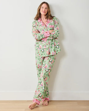 A woman stands barefoot against a white wall, smiling gently with her arms crossed. She wears the PRINTFRESH - HOLIDAY HENS LONG PAJAMA SET by PRINTFRESH, featuring light green organic cotton with a colorful floral and bird pattern and pink trim.