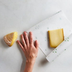 A hand holds a sheet of FORMATICUM - ZERO CHEESE STORAGE PAPER with green illustrations, next to a brown FORMATICUM box. The partially unrolled cheese storage paper is ideal for keeping cheese fresh at home.