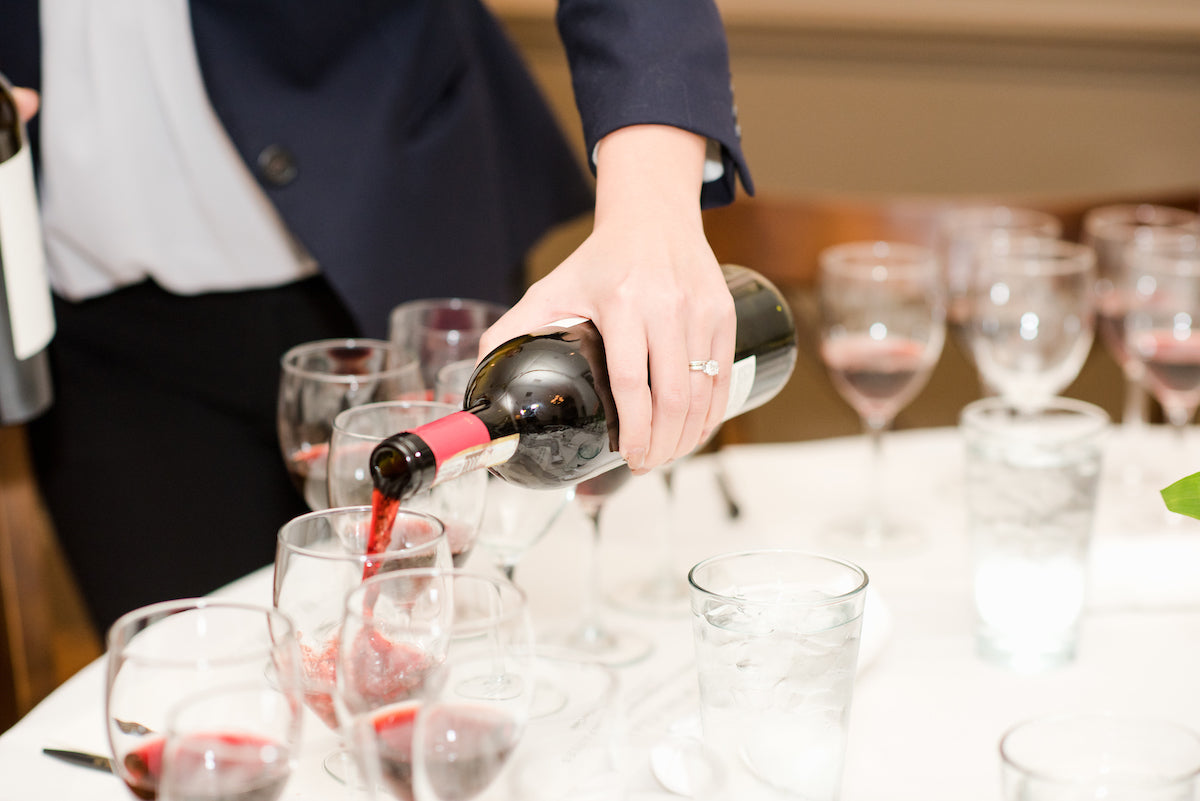 A person in a suit jacket pours red wine from a bottle into a glass on a table set with multiple wine glasses and water glasses.