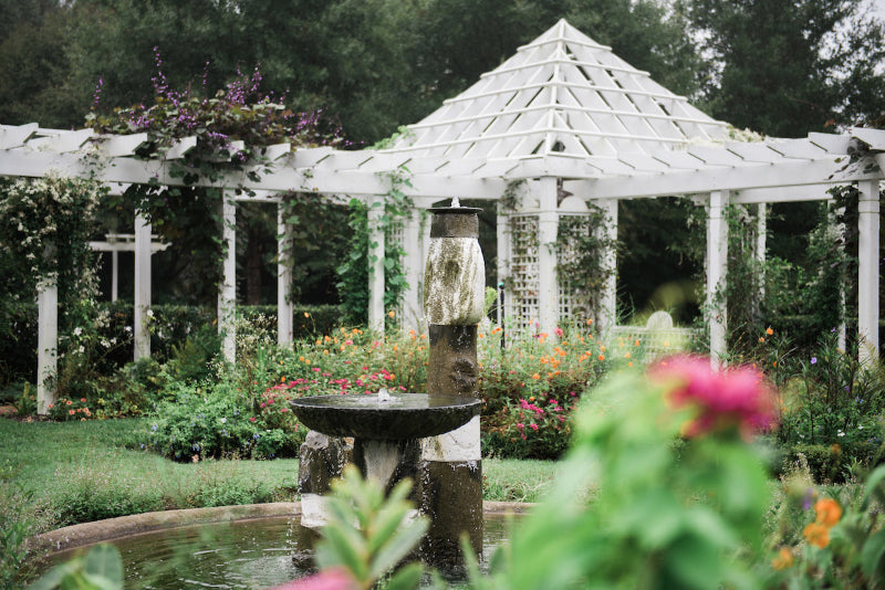 A stone fountain stands in a lush garden filled with flowers, with a white pergola and gazebo surrounded by greenery in the background.