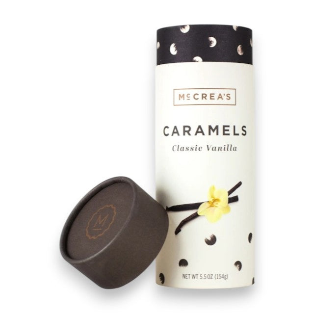 A cylindrical box of McCREA’S CANDIES - CLASSIC VANILLA CARAMELS features an elegant black and white design adorned with illustrations of vanilla flowers and pods, with the lid resting against the container. These gourmet caramels are gluten-free and have a net weight of 5.5 oz (154g).