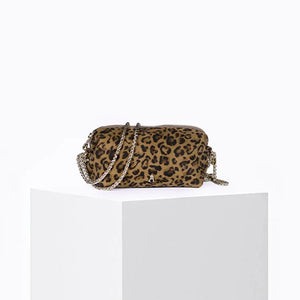 The CRAIE STUDIO - NUAGE LEO SEPIA MINI BAG, featuring a leopard print design, gold zipper, and an extended red strap, is showcased on a white pedestal against a simple backdrop.