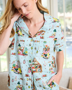 A person stands barefoot against a white wall, smiling and wearing the PRINTFRESH - READING NOOK PAJAMA SHORT SET by PRINTFRESH—colorful, organic cotton pajamas ideal for relaxing in your favorite reading nook.