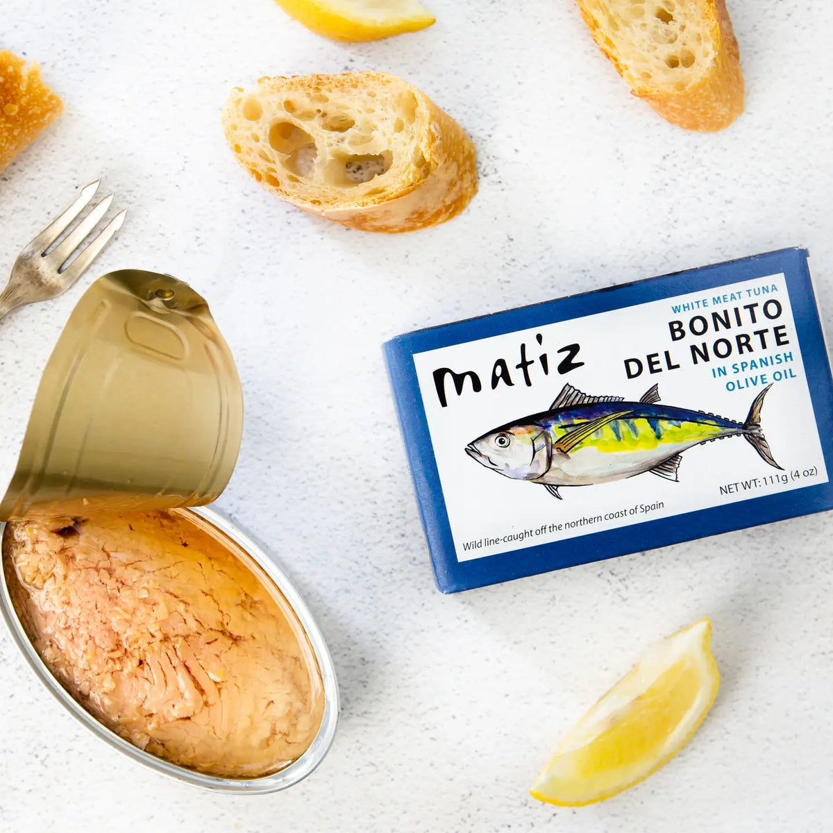 An opened can of MATIZ ESPANA - BONITO DEL NORTE IN SPANISH OLIVE OIL, in a blue box, is displayed with slices of baguette, lemon wedges, and a fork on a white surface.