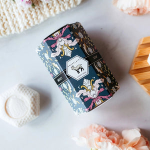 A decorative BAUDELAIRE - GOATS MILK HONEY SOAP 1.4 OZ QUARTET box sits beside a stack of four pale pink, hexagon-shaped soap bars. The floral-patterned box features an illustration of a goat, drawing attention to this gentle triple-milled soap from BAUDELAIRE, crafted with nourishing goat milk and natural honey.