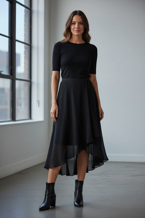 A woman stands indoors by large windows, showcasing a full-length silhouette in the ELEH Asymmetrical Skirt in Black, paired with a black short-sleeve top and black ankle boots.