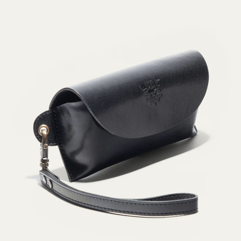 The WILL LEATHER GOODS - EYEGLASS CASE IN BLACK is a handcrafted Italian leather case with a flap closure, embossed logo on the front, and an attached wrist strap on the left, shown against a plain light background.