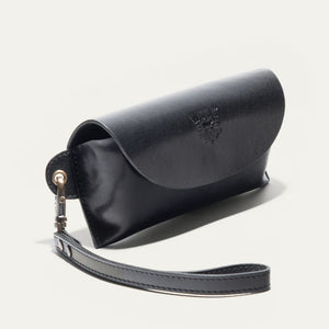 The WILL LEATHER GOODS - EYEGLASS CASE IN BLACK is a handcrafted Italian leather case with a flap closure, embossed logo on the front, and an attached wrist strap on the left, shown against a plain light background.