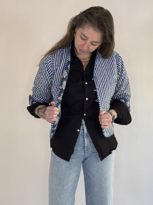 A woman with visible forearm tattoos smiles slightly against a plain background, wearing the FARYLINA ZOIE Reversible Quilted Jacket over a denim shirt, white pants, and a tan belt.