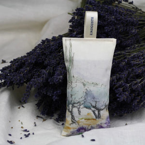 A rectangular L’AUGUSTE PROVENCE Lavender Sachet from Provence, featuring a scenic design and labeled fabric loop, stands upright on a white background with scattered Provence lavender buds in front.