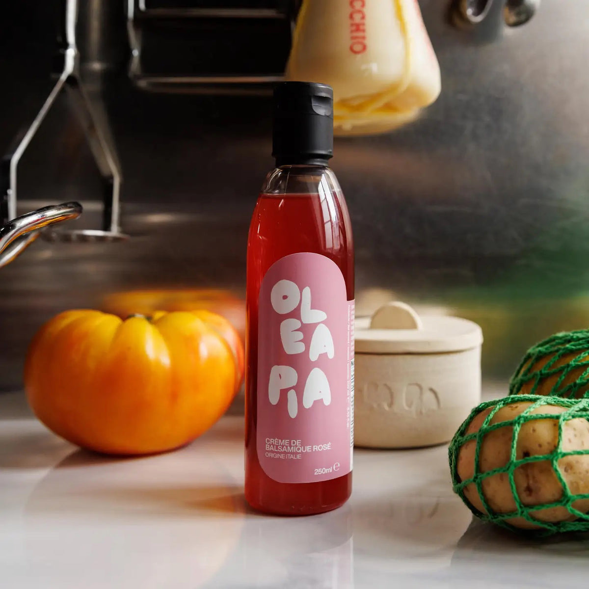 A bottle of Olea Pia red sauce sits on a kitchen countertop, giving off fruity aromas. It is surrounded by a yellow tomato, a netted melon, and a small ceramic container of Olea Pia - Pink Balsamic Vinegar Cream. In the background, the metallic backsplash and various kitchen utensils are visible.
