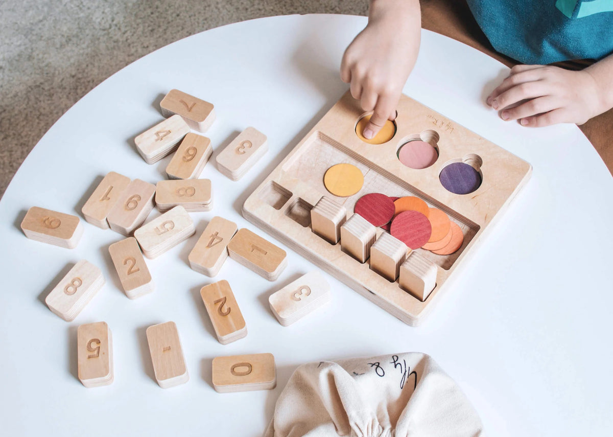 The "LITTLE NUMBERS GAME SET" by LILY &amp; RIVER is a wooden educational toy influenced by Montessori principles. It features number blocks and colored disks on a white surface. The blocks display a simple math equation (2 + 2 = 4), functioning like a children's calculator. In the background, there is a light-colored fabric bag with the text "Lily &amp; River" partially visible.
