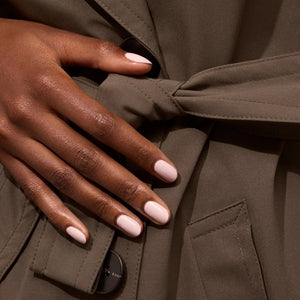 A hand with pale pink polished nails holds a bottle of MANUCURIST INC GREEN™ Nail Polish in Blossom against a gray knit sweater background.