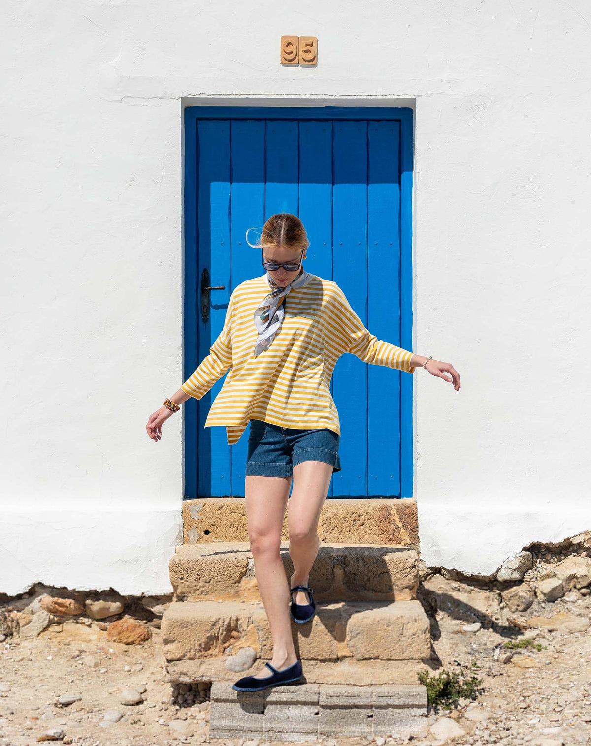 Wearing the MERSEA Catalina Jersey Tee in Golden Rod, a woman with 3/4 length sleeves and denim shorts steps down stone stairs in front of a vibrant blue door set within a white wall.