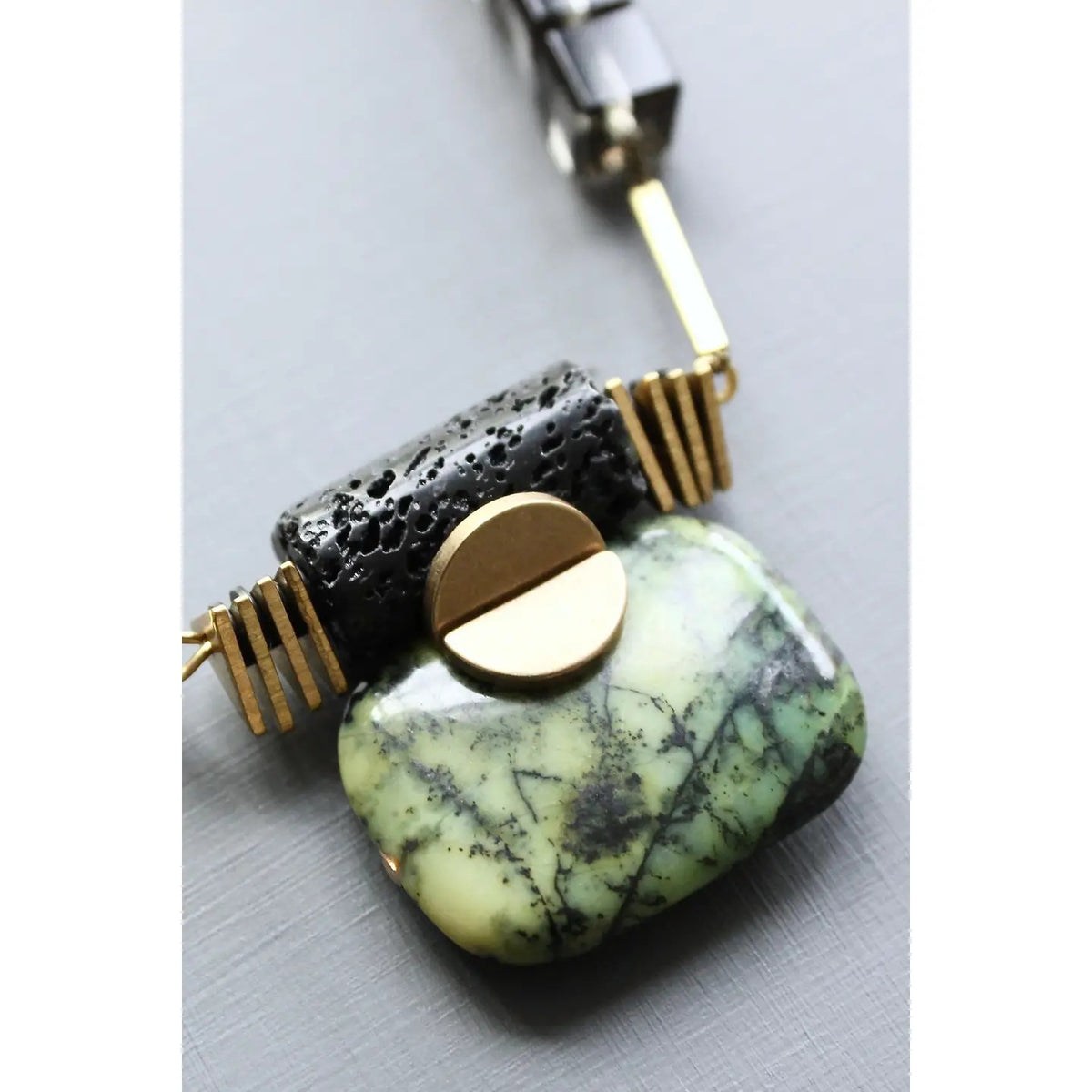 Introducing the DAVID AUBREY - JASPER ARTDECO PENDANT NECKLACE by DAVID AUBREY, featuring a gold chain adorned with dark cylindrical beads and a striking green marbled pendant evocative of green jasper, highlighted by elegant gold and silver accents. Displayed against a light gray background.