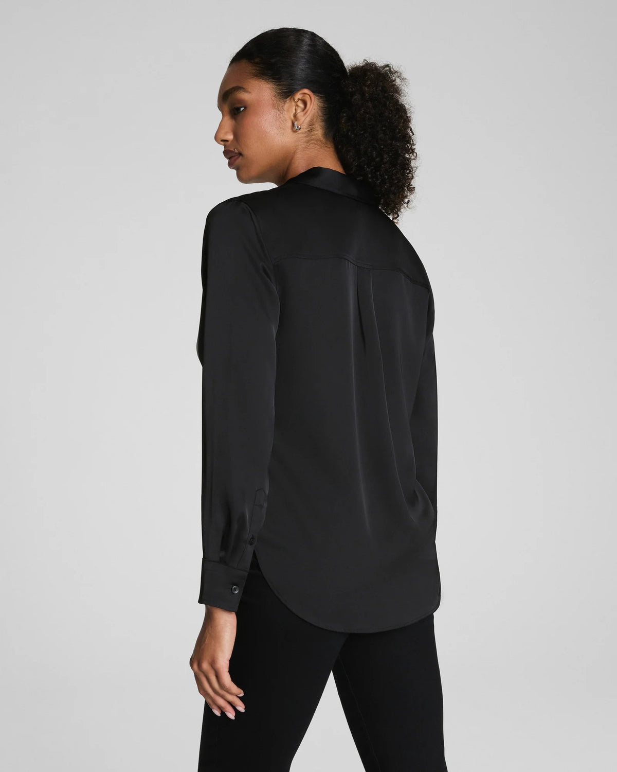 A woman with curly hair pulled back wears the SPANX Silky Smooth Button Down Shirt in black, paired with black pants. She stands facing away, looking over her shoulder against a plain light gray background.