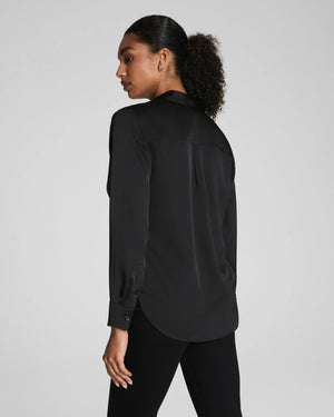 A woman with curly hair pulled back wears the SPANX Silky Smooth Button Down Shirt in black, paired with black pants. She stands facing away, looking over her shoulder against a plain light gray background.