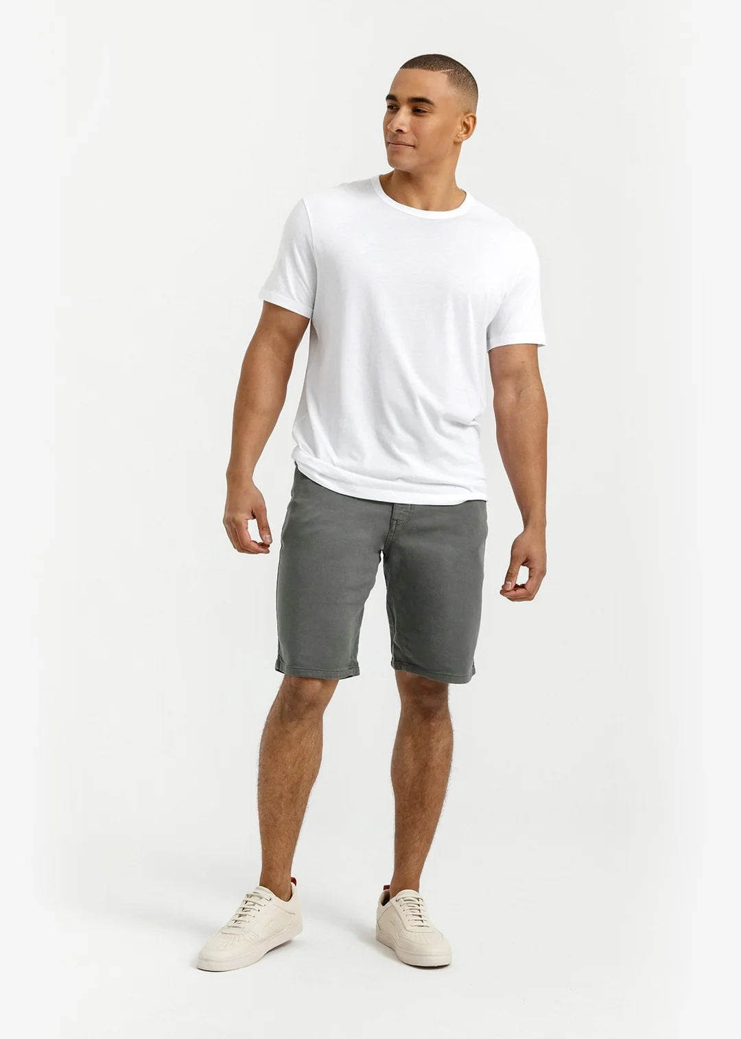 A person stands against a plain light background, wearing a white T-shirt, DUER No Sweat Relaxed Shorts in Gull made from stretch fabric, and white sneakers, with one hand in their pocket.