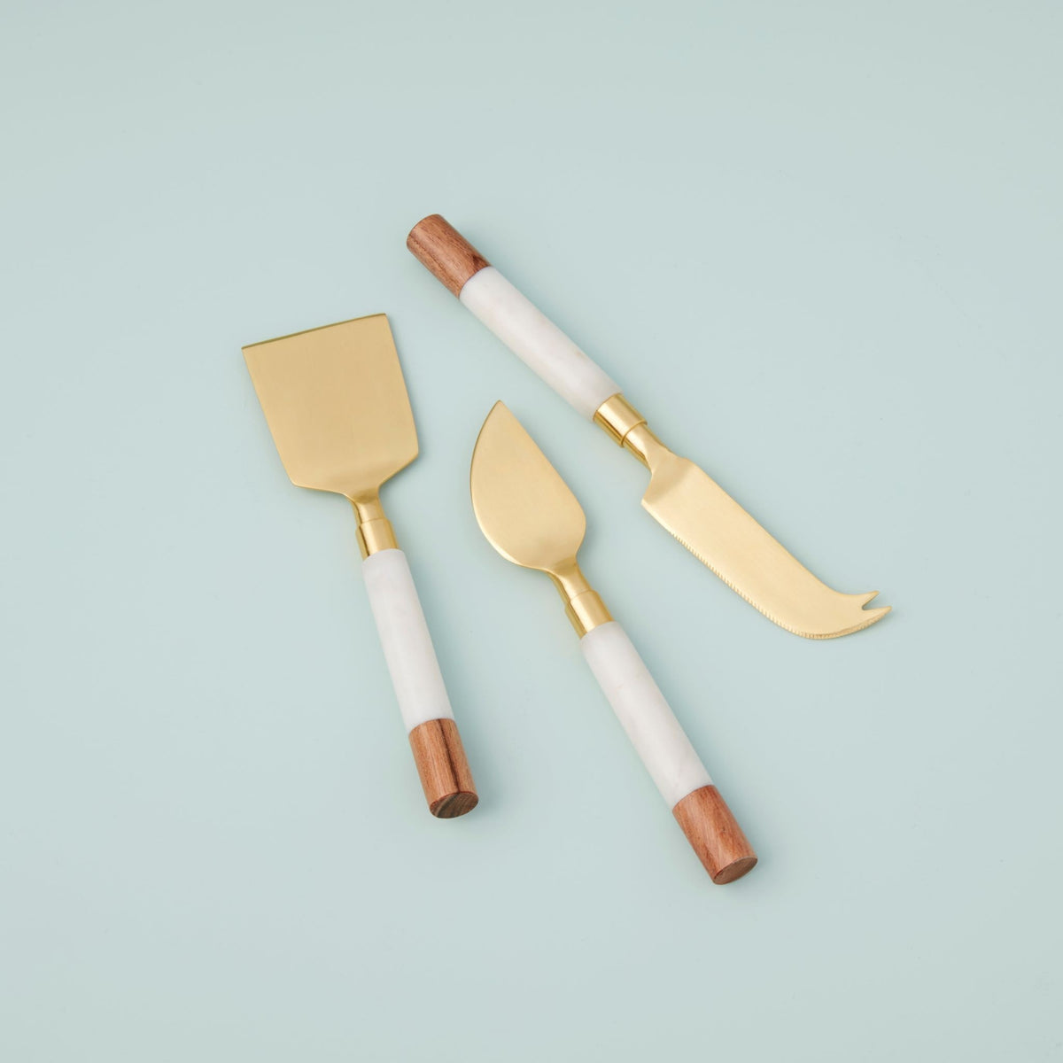 The BE HOME - GARNET CHEESE SET by BE HOME, INC features three elegant cheese knives with gold-toned blades and white handles accented with wood—perfect for elevating your charcuterie spread in both style and function.