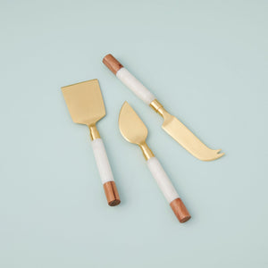 The BE HOME - GARNET CHEESE SET by BE HOME, INC features three elegant cheese knives with gold-toned blades and white handles accented with wood—perfect for elevating your charcuterie spread in both style and function.