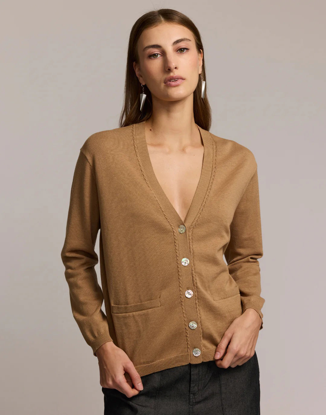 A woman with straight brown hair wears the HINSON WU CASSIE Long Sleeve Cotton Silk Cardigan in camel, styled with black pants. She poses confidently, one hand to her chin, against a neutral backdrop.