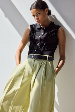 A person stands against a white wall in sunlight, wearing the HOPE FOR FLOWERS Peter Pan Sleeveless Top in black by HOPE FOR FLOWERS, paired with high-waisted beige wide-leg pants and a black belt, hands in their pockets and a neutral expression.