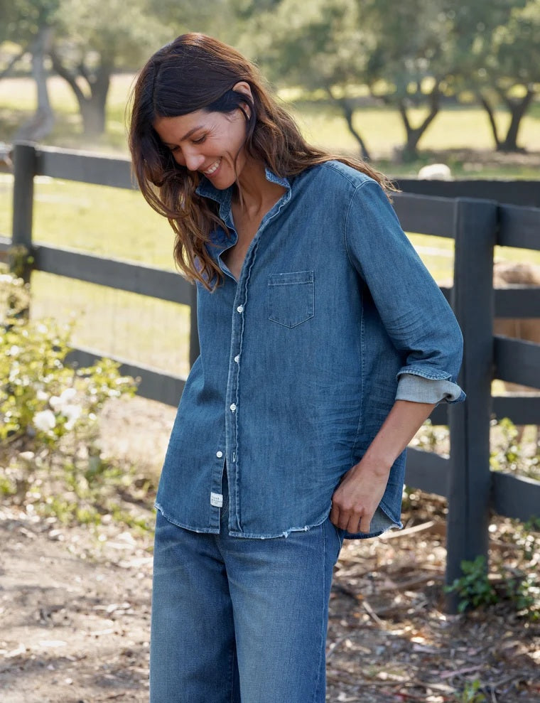 A person with long brown hair stands outdoors, facing away, next to a black wooden fence with trees and grass in the background, wearing the FRANK & EILEEN EILEEN Relaxed Button Up Shirt in Famous Denim Distressed Indigo Wash.